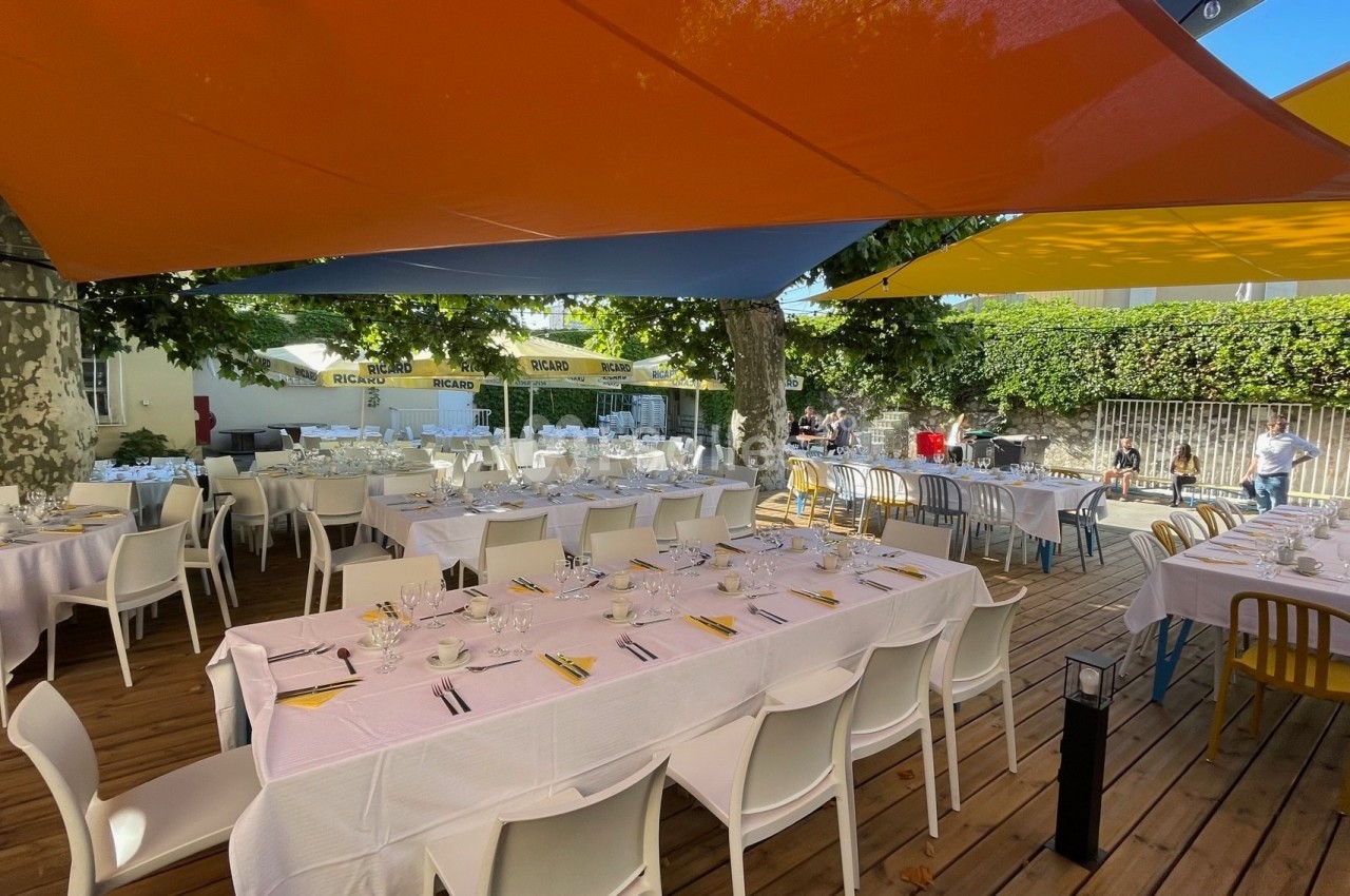Tables dressées avec nappes blanches sous des voiles d'ombrage colorées, dans un espace extérieur entouré de verdure.