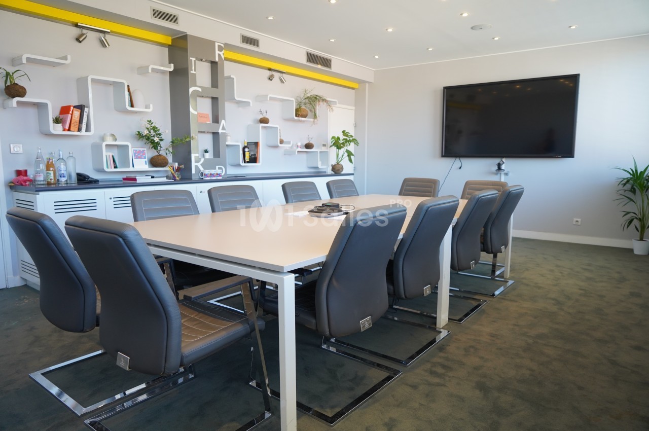 Salle de réunion moderne avec table rectangulaire, chaises grises, écran mural et étagères décorées.