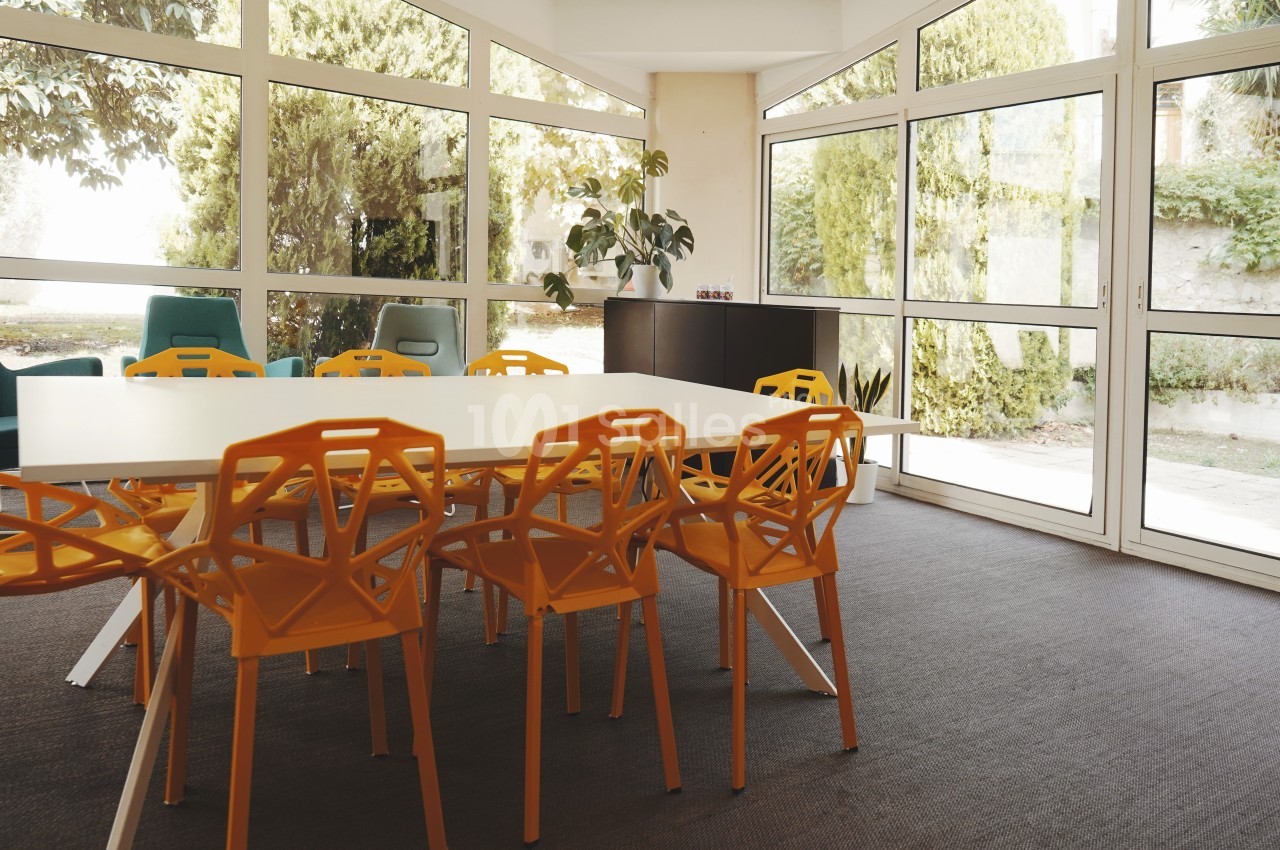 Salle de réunion lumineuse avec table blanche, chaises orange et grandes baies vitrées donnant sur un jardin.