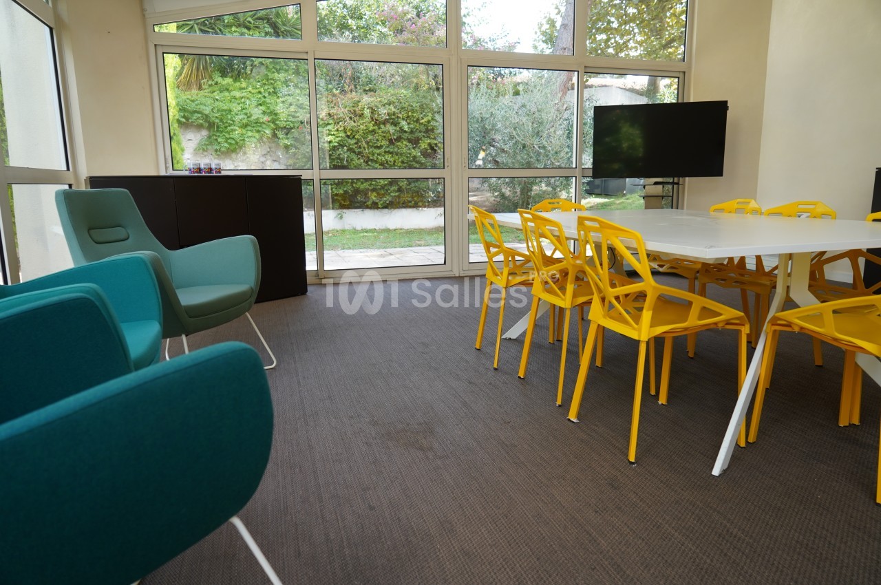 Salle de réunion lumineuse avec chaises jaunes, table blanche, fauteuils bleus et vue sur un jardin à travers de grandes…