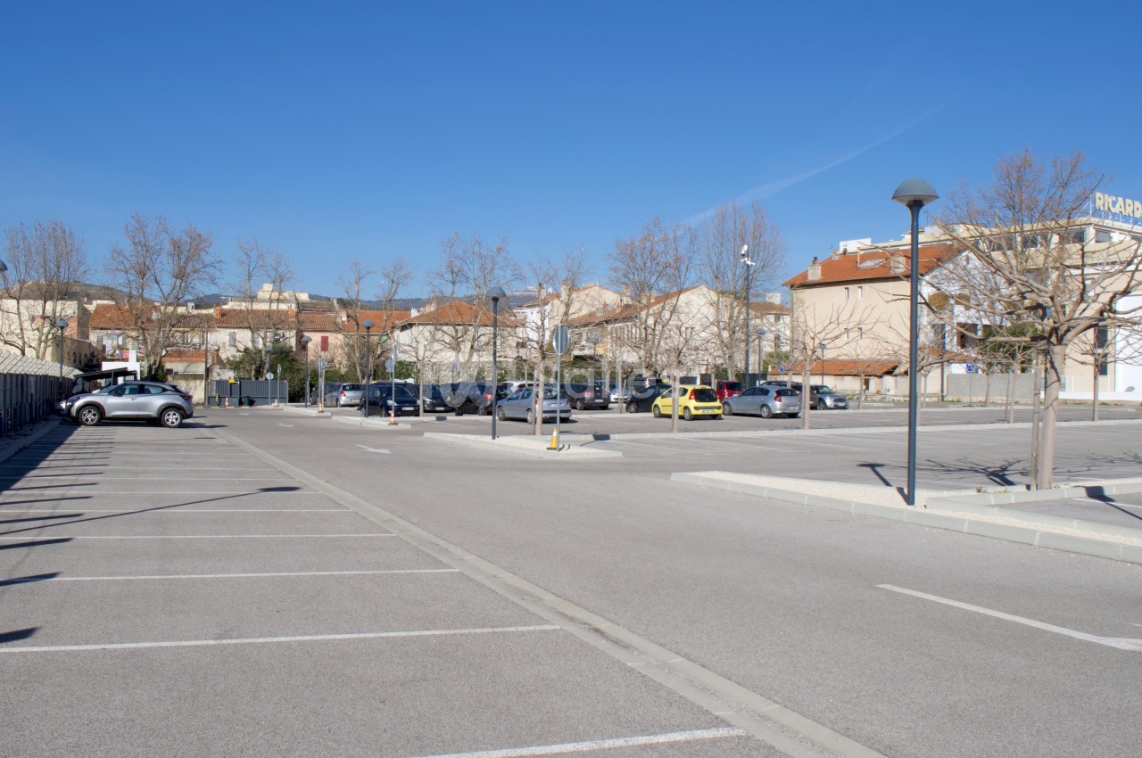 Parking extérieur vide en grande partie, bordé d'arbres sans feuilles, avec quelques bâtiments en arrière-plan.