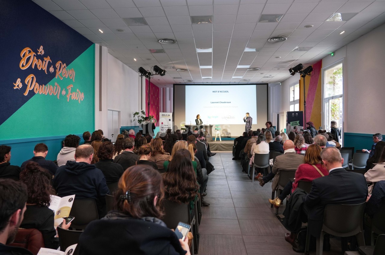 Salle de conférence avec un public attentif, un intervenant sur scène et une présentation projetée à l'écran.