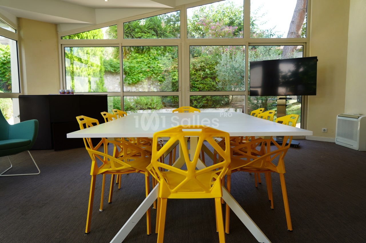 Salle de réunion lumineuse avec table blanche, chaises jaunes au design moderne et écran noir devant des baies vitrées.