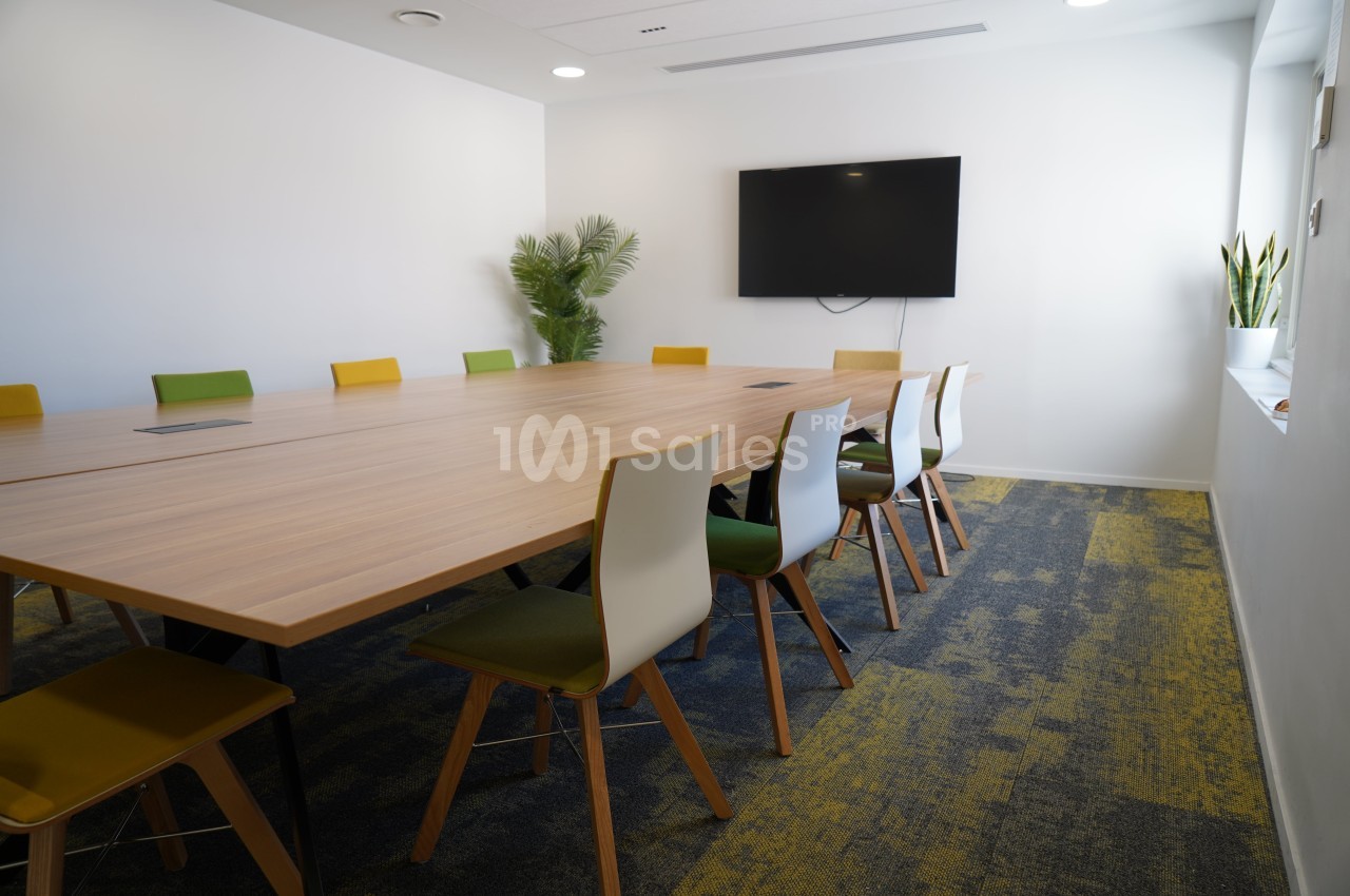 Salle de réunion lumineuse avec une grande table en bois, des chaises colorées et un écran mural.