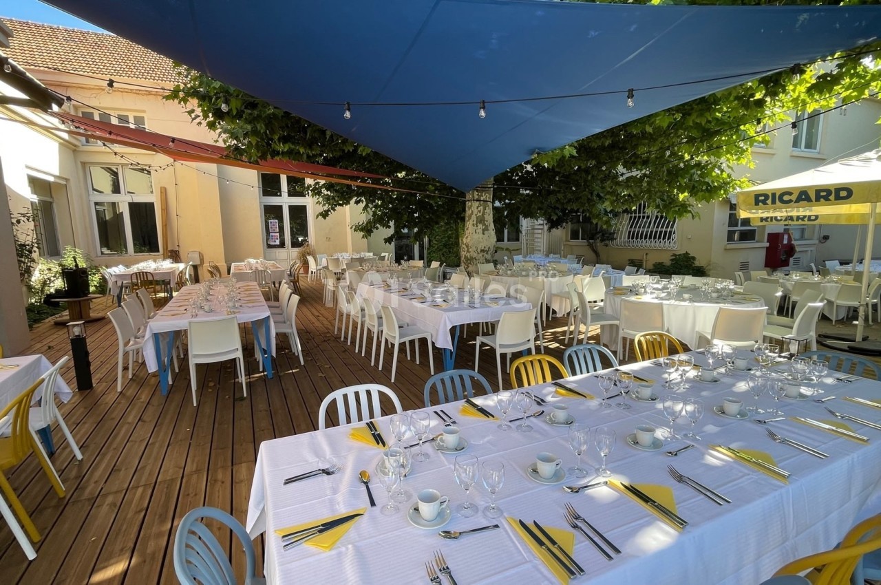 Terrasse en bois aménagée avec des tables dressées sous des voiles d'ombrage bleues et des arbres.
