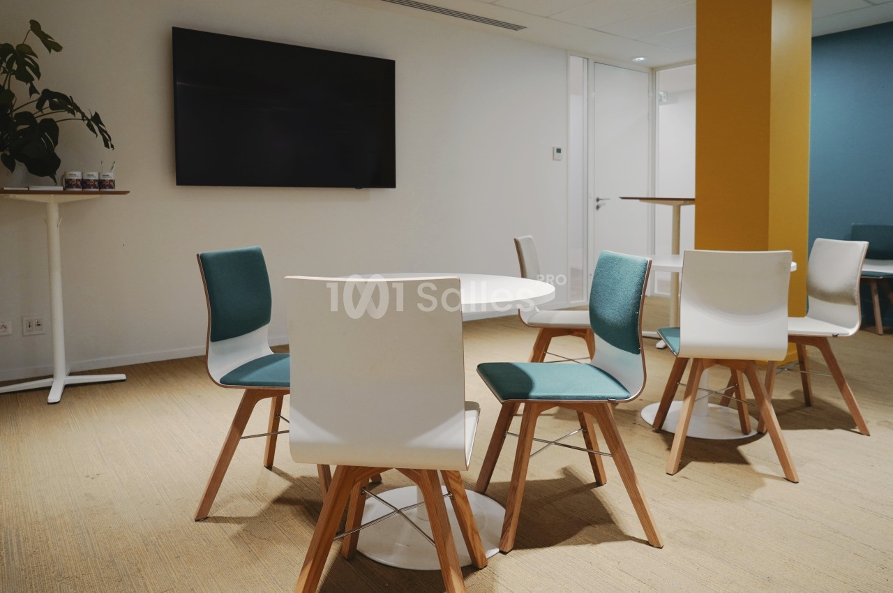 Salle de réunion avec chaises blanches et vertes autour de tables rondes, écran mural et sol en moquette beige.