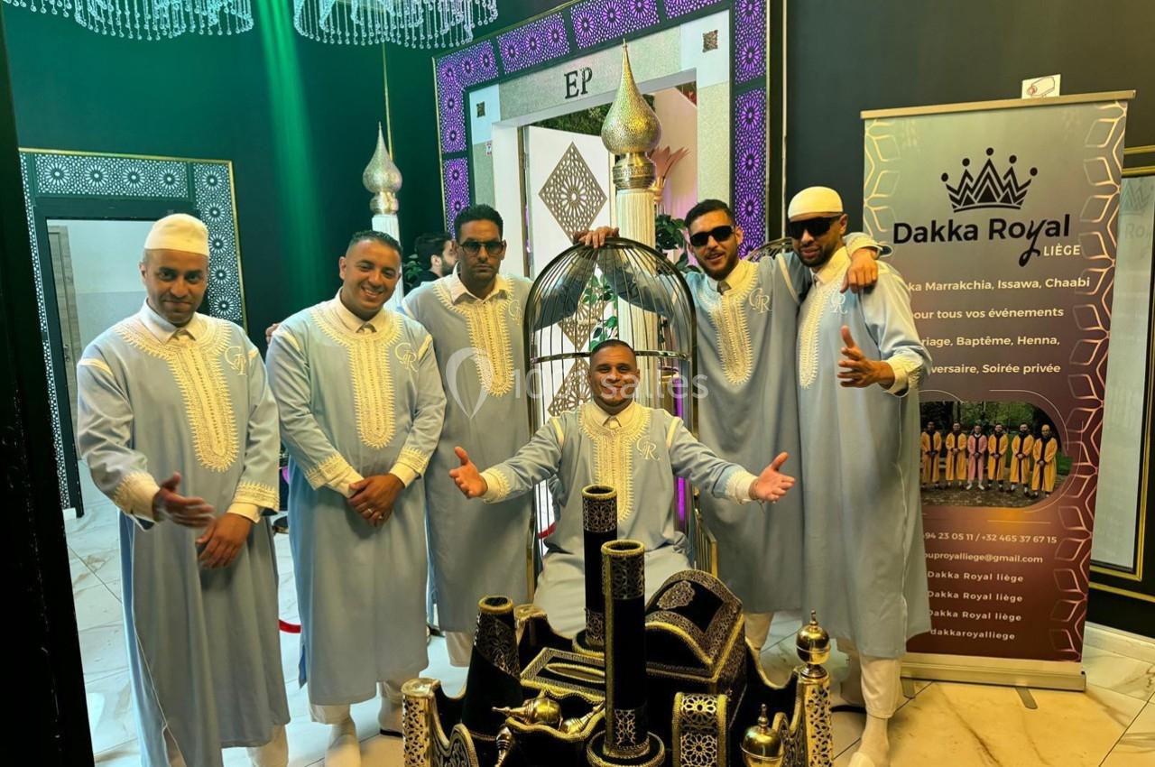 Un groupe d'hommes en tenues traditionnelles pose devant une décoration orientale et une affiche promotionnelle.