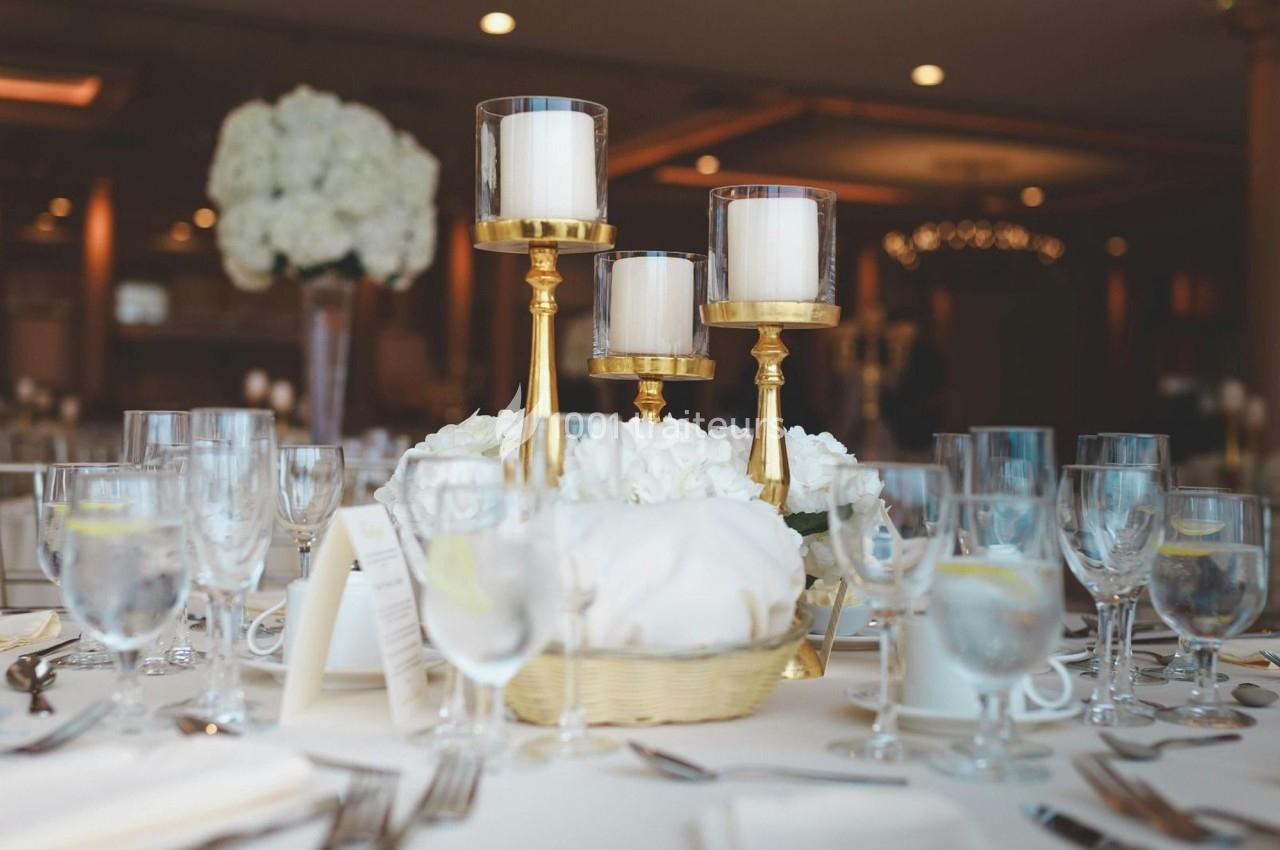 Table élégamment dressée avec chandeliers dorés, verres, assiettes et centre de table floral blanc.