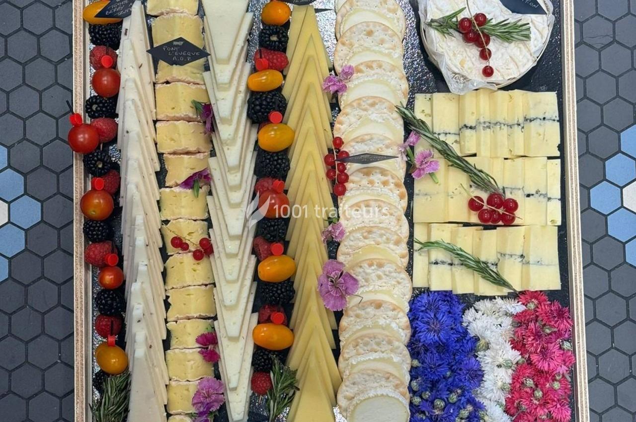 Plateau de fromages variés avec fruits, fleurs comestibles et décorations, présenté sur un fond de carreaux hexagonaux.