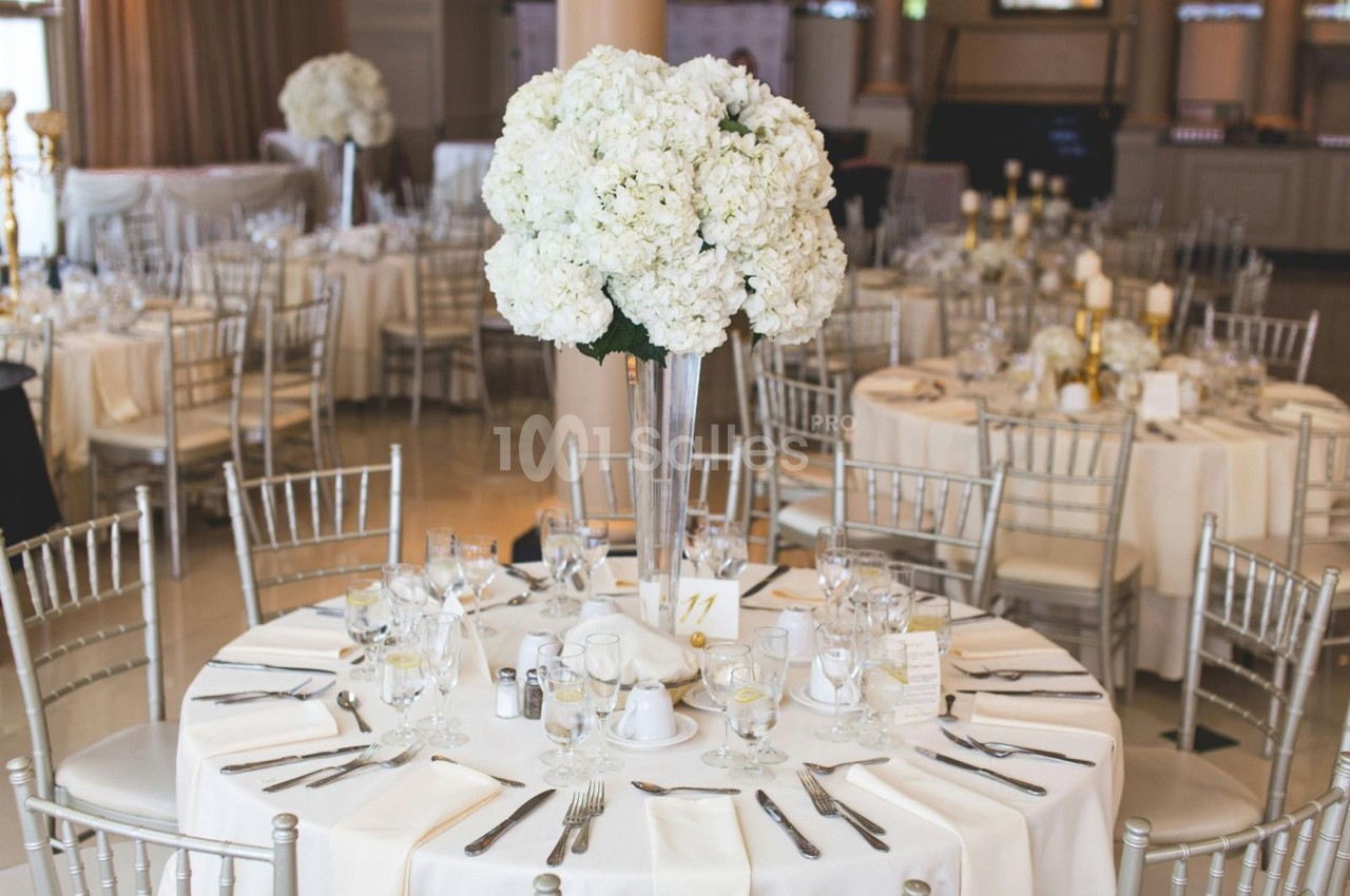 Salle de réception avec tables rondes dressées, nappes blanches, chaises argentées et centres de table fleuris.