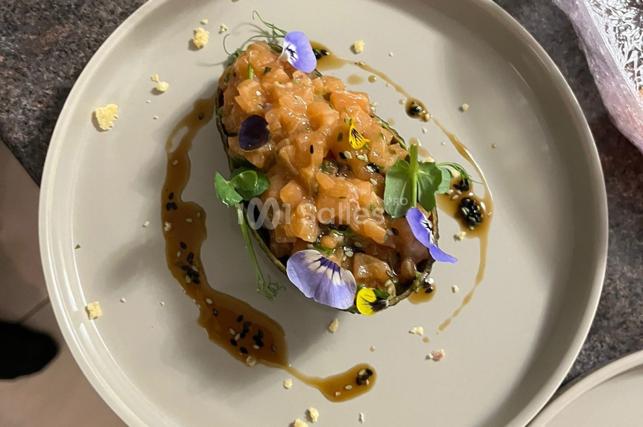 Tartare de saumon présenté dans une coque d'avocat, décoré de fleurs comestibles et entouré de sauce.