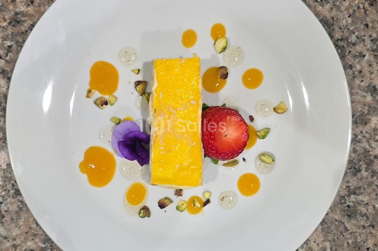 Dessert rectangulaire jaune avec fraise, pistaches, fleurs comestibles et sauces colorées sur une assiette blanche.