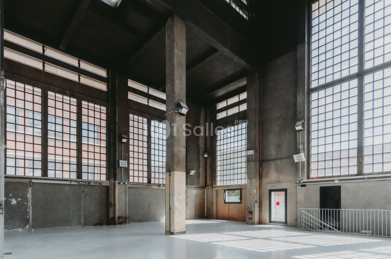 Intérieur d'un grand bâtiment industriel avec de hauts plafonds, des colonnes en béton et de larges fenêtres quadrillées.