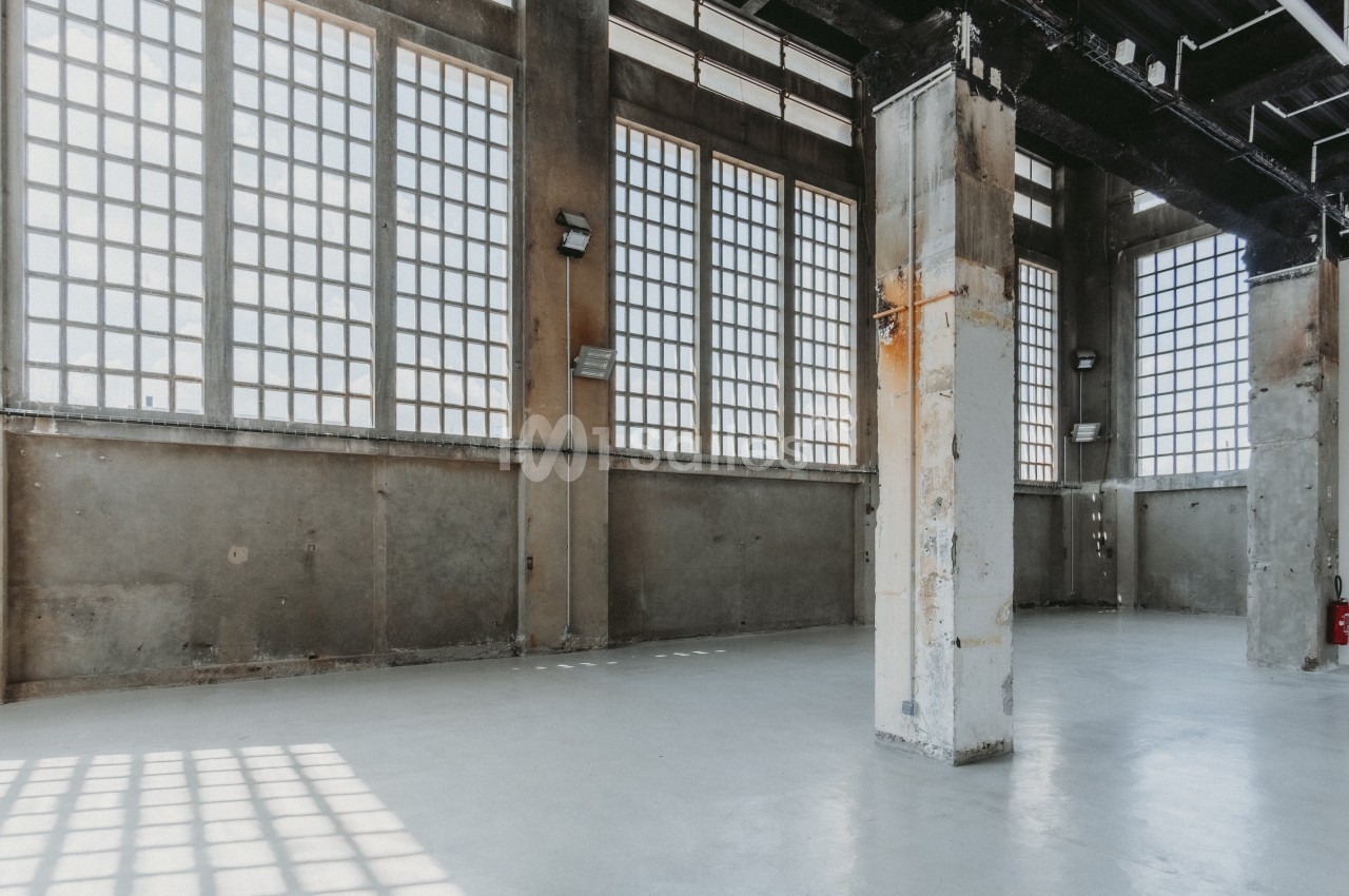 Intérieur d'un grand bâtiment industriel vide avec de hauts murs en béton et des fenêtres à carreaux.