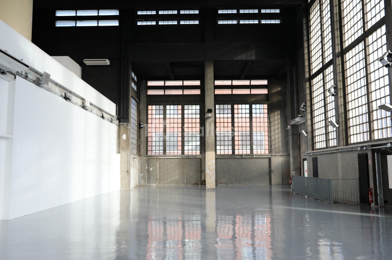 Intérieur d'un grand espace industriel avec sol brillant, murs en béton et grandes fenêtres rectangulaires.