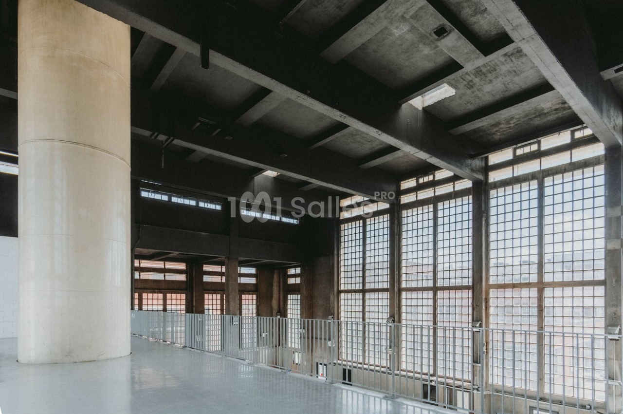 Intérieur d'un bâtiment industriel avec de grandes fenêtres à carreaux, un plafond en béton et une rampe métallique.