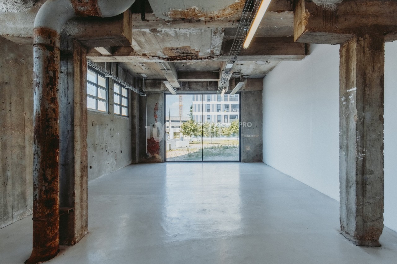 Intérieur d'un espace industriel vide avec murs en béton brut, tuyaux apparents et grande baie vitrée donnant sur l…