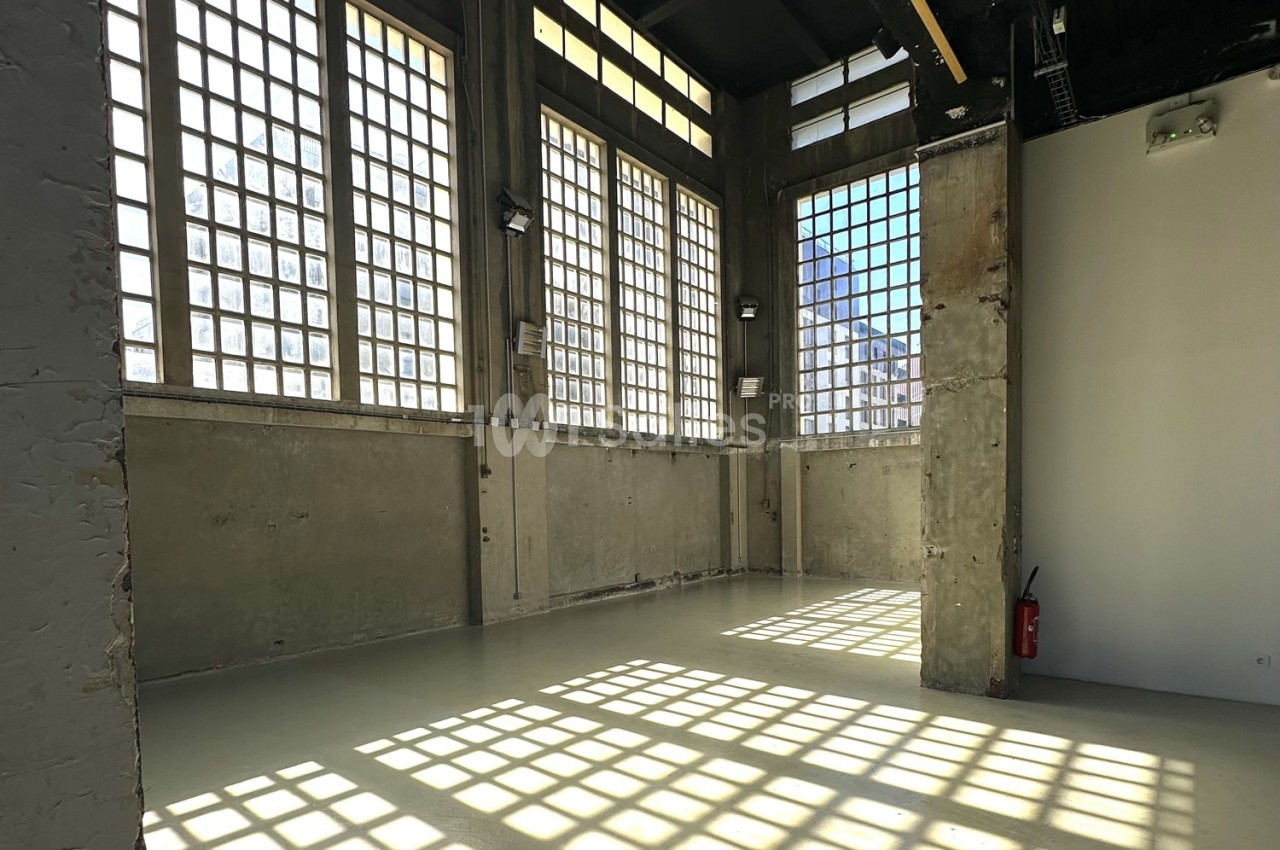 Intérieur d'une pièce lumineuse avec de grandes fenêtres à carreaux, murs en béton brut et un extincteur au fond.
