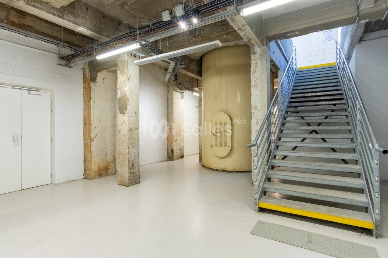 Escalier métallique montant à droite, murs en béton brut et réservoir cylindrique beige dans un espace industriel.