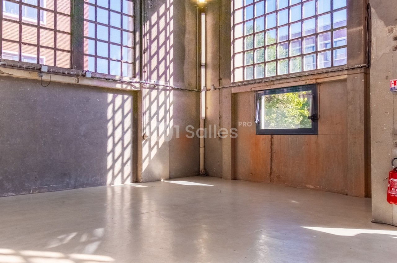 Intérieur d'un espace industriel vide avec sol en béton, grandes fenêtres à carreaux et lumière naturelle.