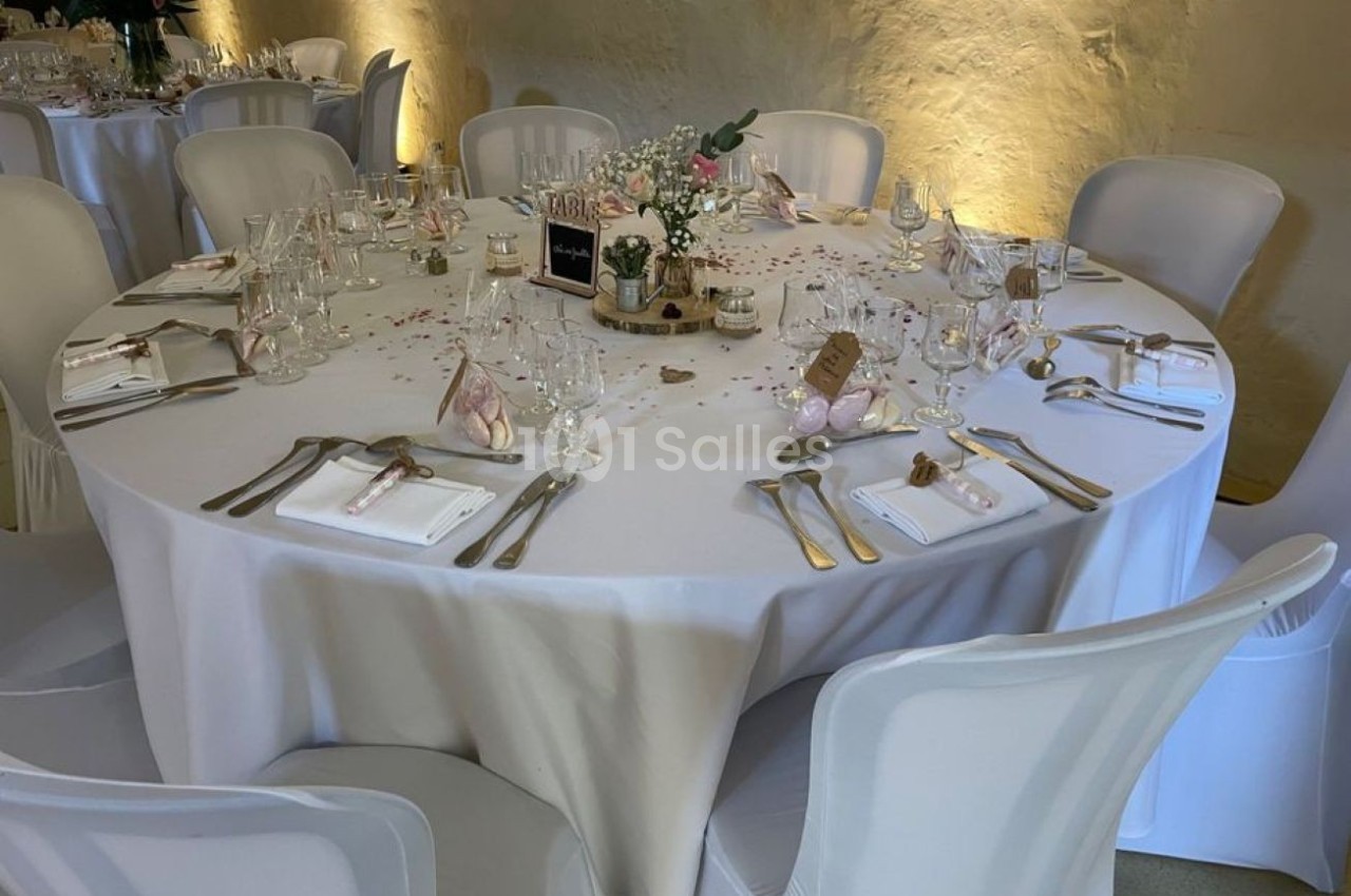 Table ronde élégamment dressée avec nappes blanches, couverts, verres et décorations florales dans une salle éclairée.