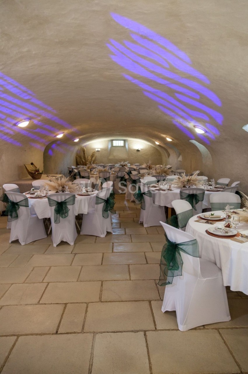 Salle voûtée décorée pour un événement, avec des tables rondes dressées et des chaises ornées de nœuds verts.