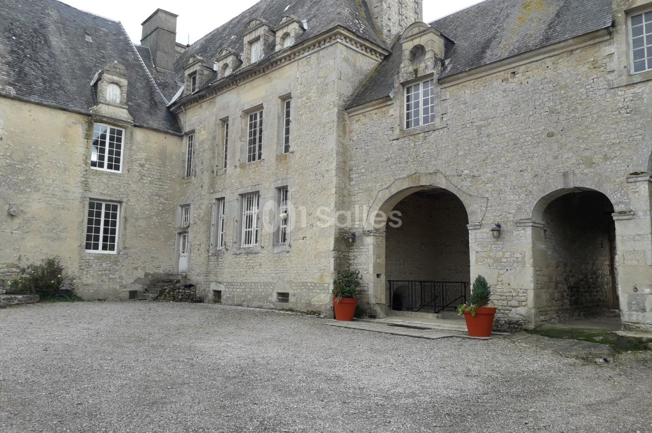 Cour intérieure d'un bâtiment en pierre de style ancien, avec des arches, des fenêtres et deux pots de fleurs rouges.