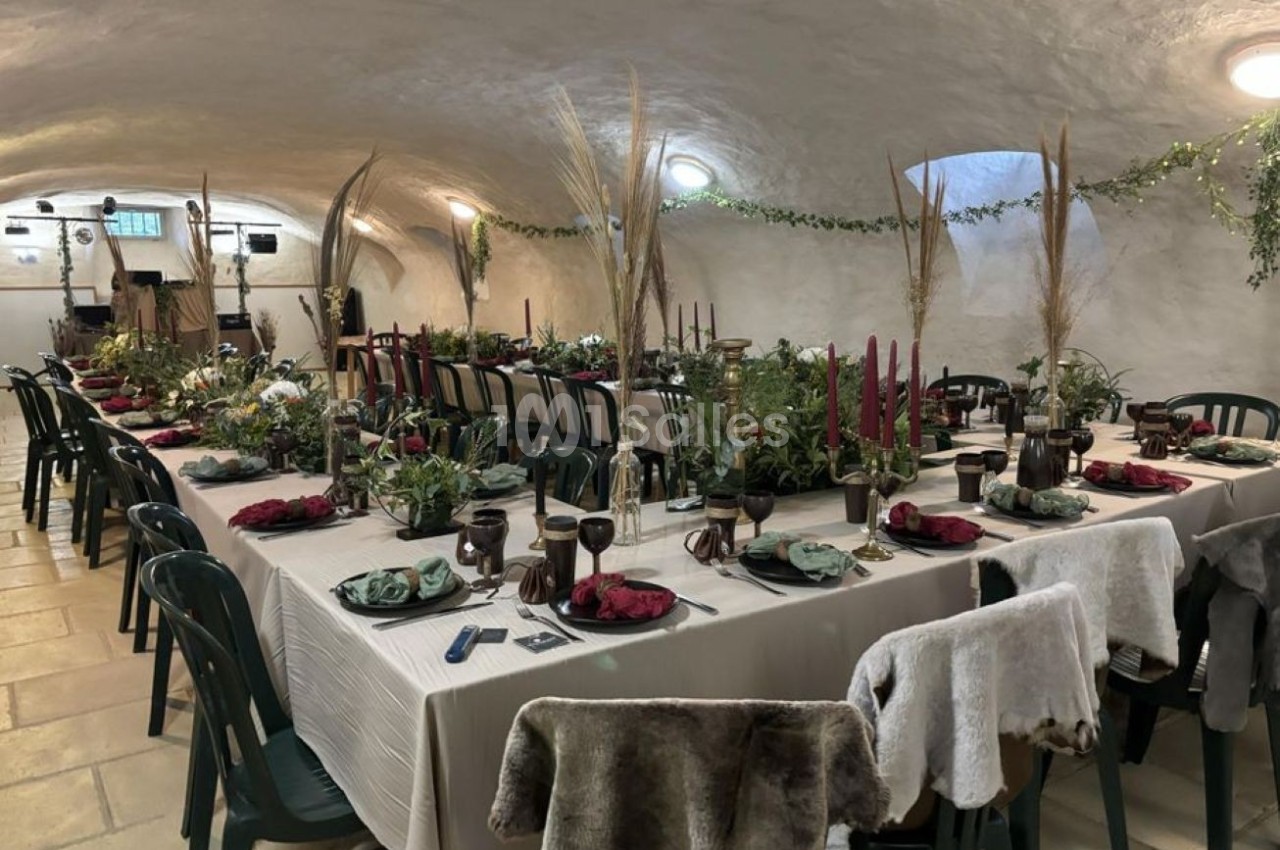 Salle voûtée aménagée pour un repas, avec longues tables décorées de bougies, plantes et serviettes colorées.