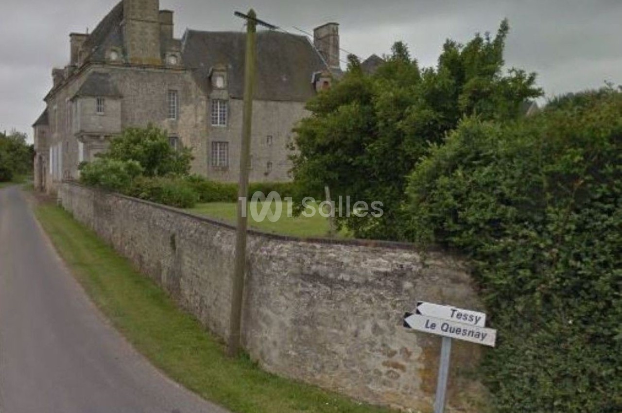 Vue d'une route de campagne bordée d'un mur en pierre, avec un bâtiment ancien en arrière-plan et un panneau indiquant…