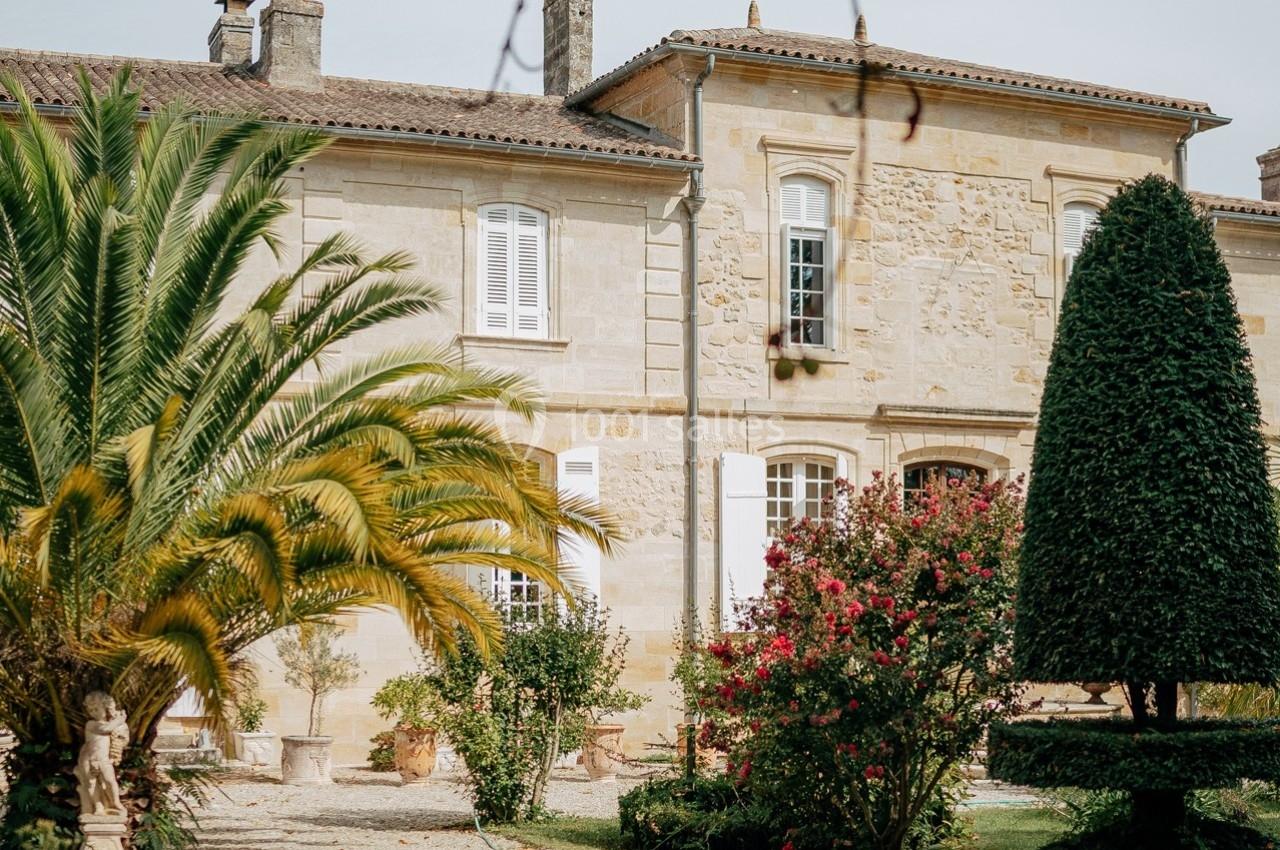 Façade d'une maison en pierre entourée de palmiers, arbustes fleuris et un jardin bien entretenu.