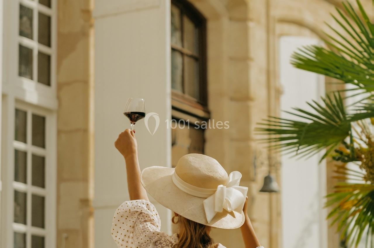 Femme de dos, vêtue d'un chapeau et d'une tenue claire, levant un verre de vin devant une façade en pierre avec volets…