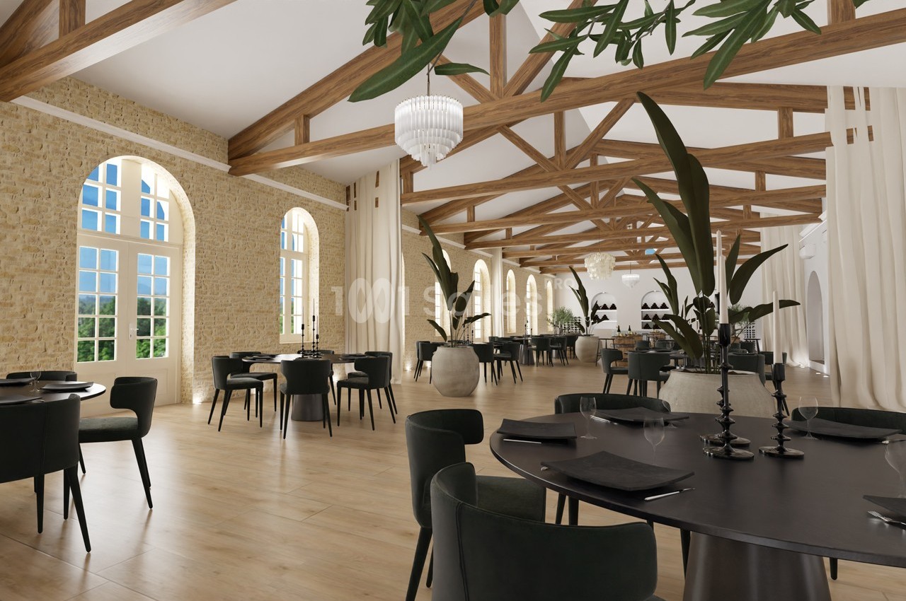 Salle de restaurant lumineuse avec poutres apparentes, grandes fenêtres, tables noires et plantes décoratives.
