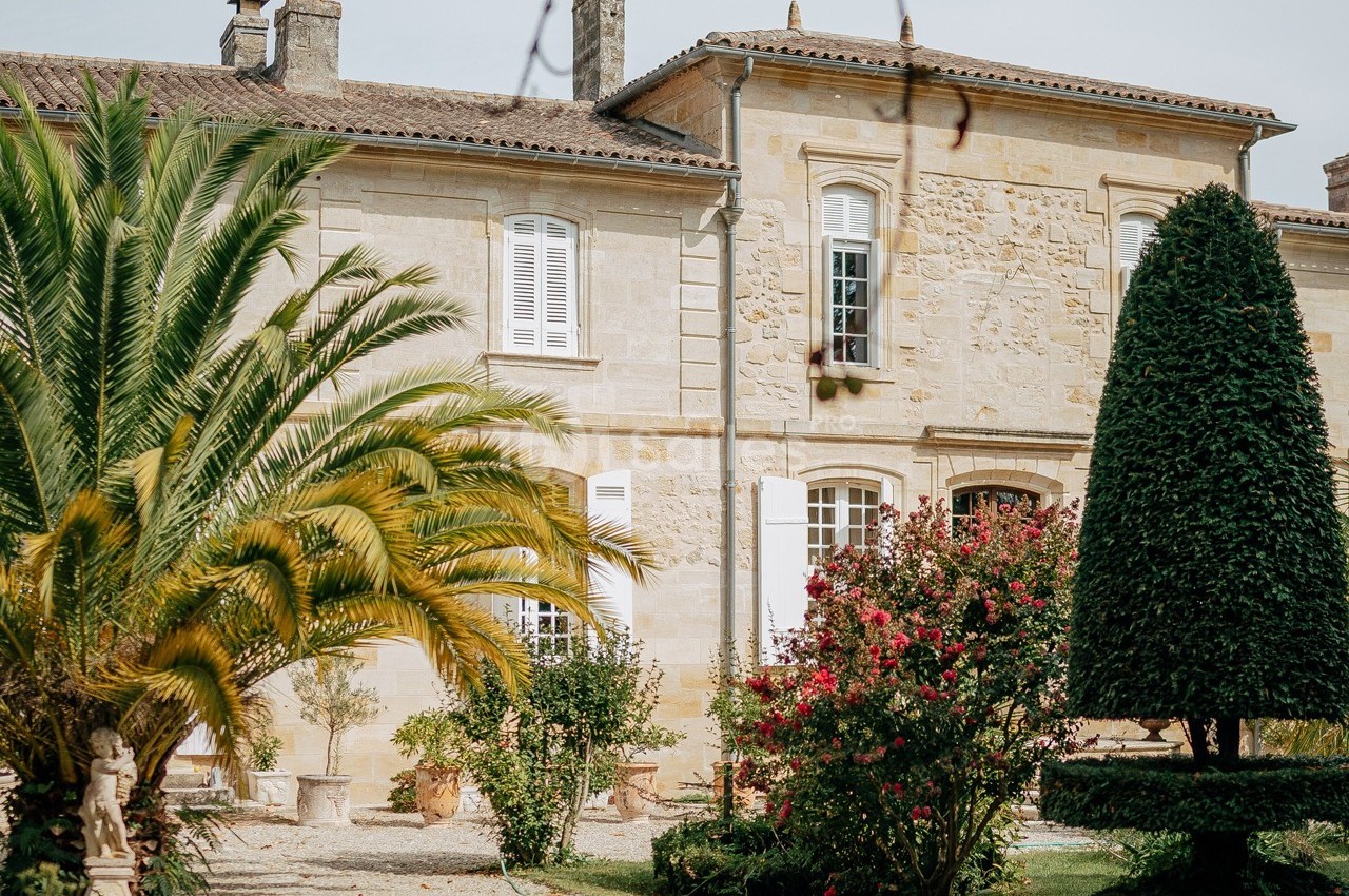 Façade d'une maison en pierre entourée de palmiers, arbustes fleuris et un jardin bien entretenu.