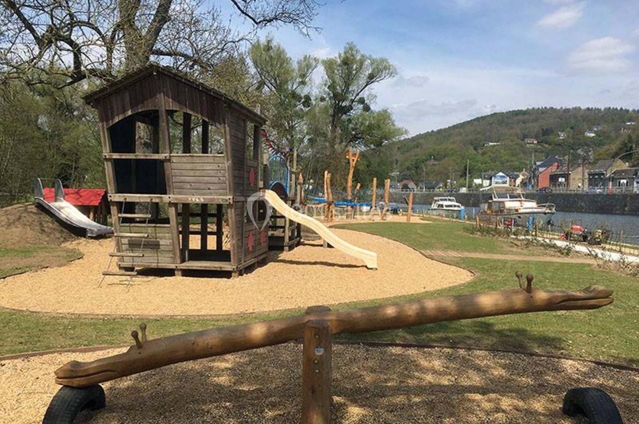 Aire de jeux extérieure avec cabane en bois, toboggans, balançoire à bascule et vue sur un paysage verdoyant.
