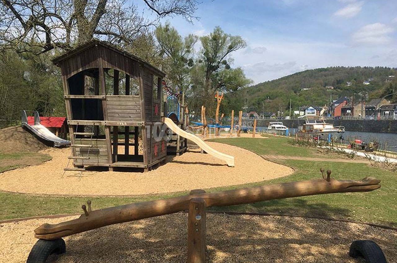 Aire de jeux extérieure avec cabane en bois, toboggans, balançoire à bascule et vue sur un paysage verdoyant.