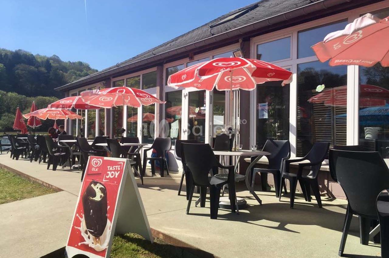 Terrasse ensoleillée avec des tables, chaises noires et parasols rouges, devant un bâtiment vitré.