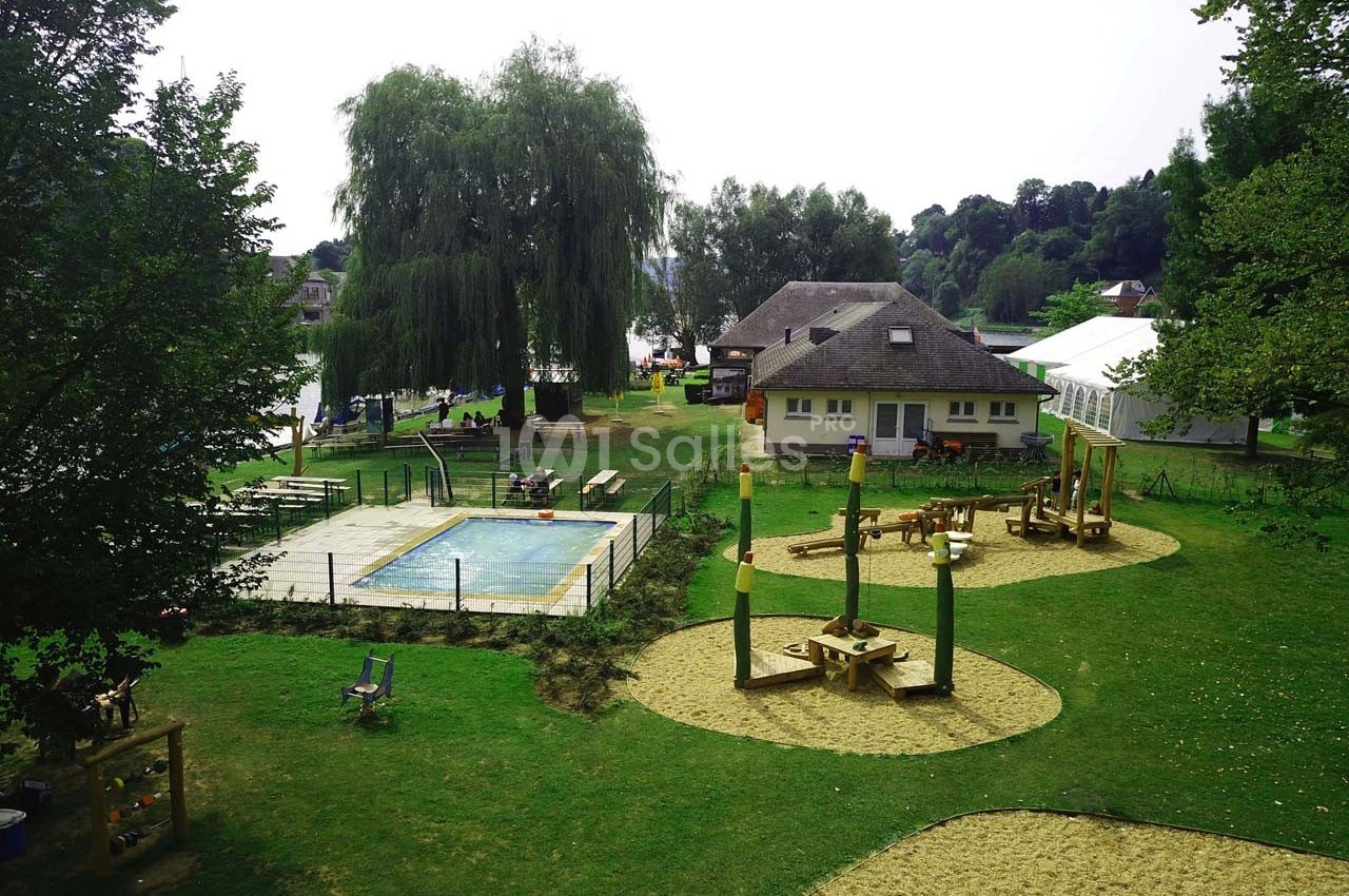 Aire de jeux extérieure avec structures en bois, piscine clôturée et espaces verts entourés d'arbres.