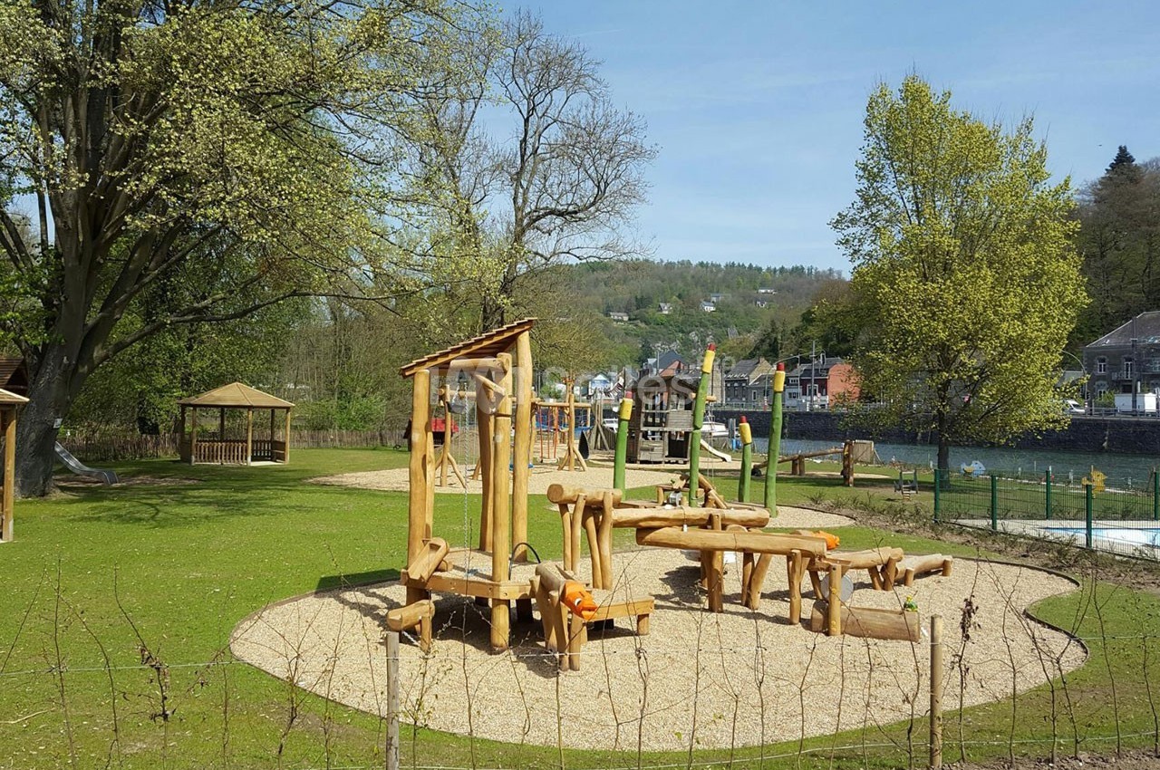 Aire de jeux en bois sur un sol sablé, entourée de verdure et située près d'une rivière et de collines.