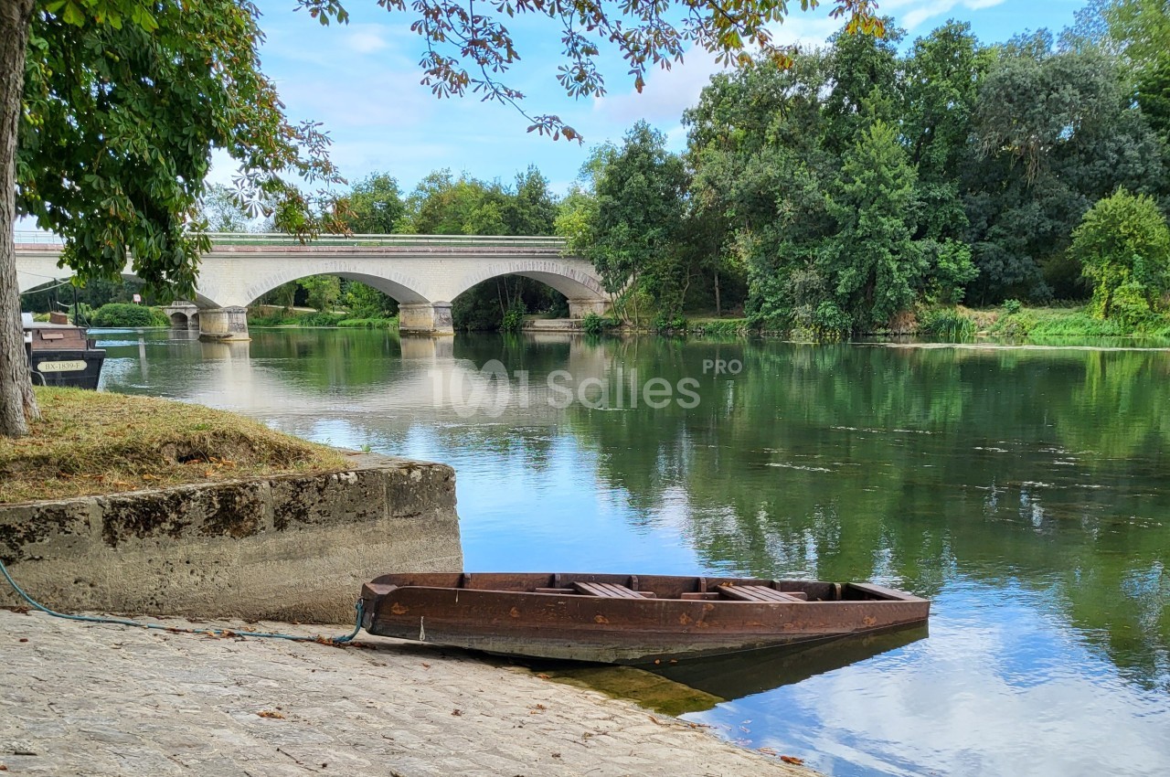 Barque en bois amarrée sur une rive pavée, près d'une rivière bordée d'arbres et d'un pont en pierre.