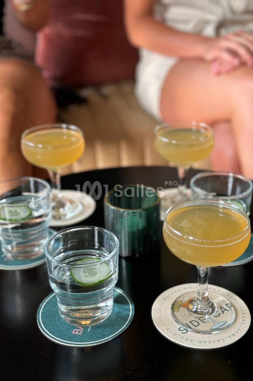 Verres de cocktails jaunes et d'eau avec rondelles de concombre posés sur une table noire, entourés de convives.