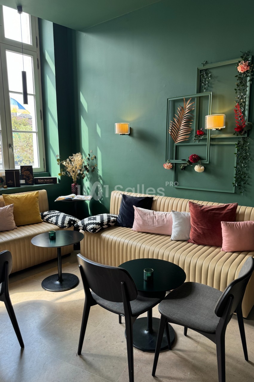 Salon cosy avec banquettes beige, coussins colorés, tables noires et décoration murale florale sur fond vert.