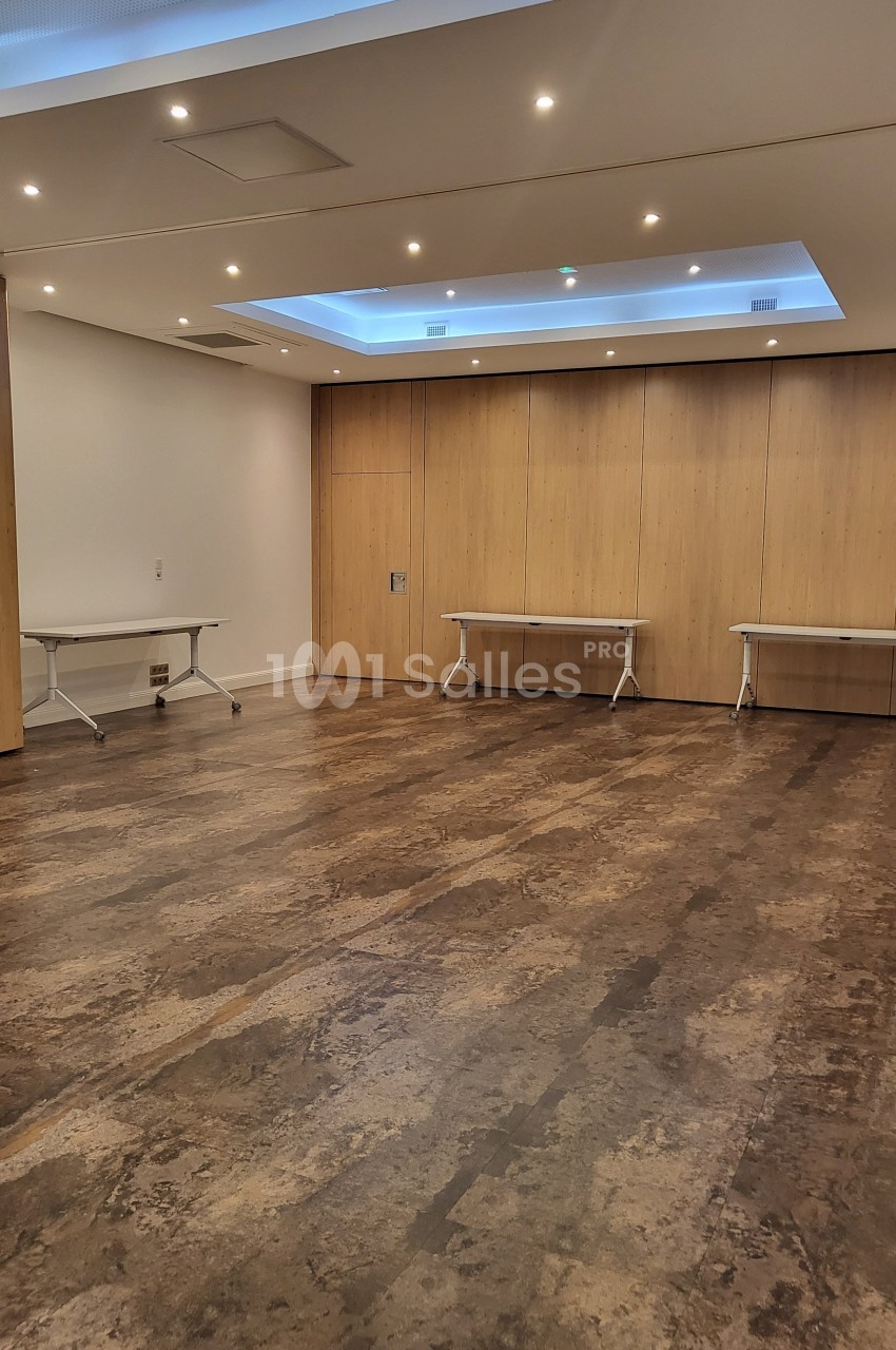 Salle vide avec parquet sombre, murs en bois clair, plafond lumineux et deux tables pliantes contre le mur.