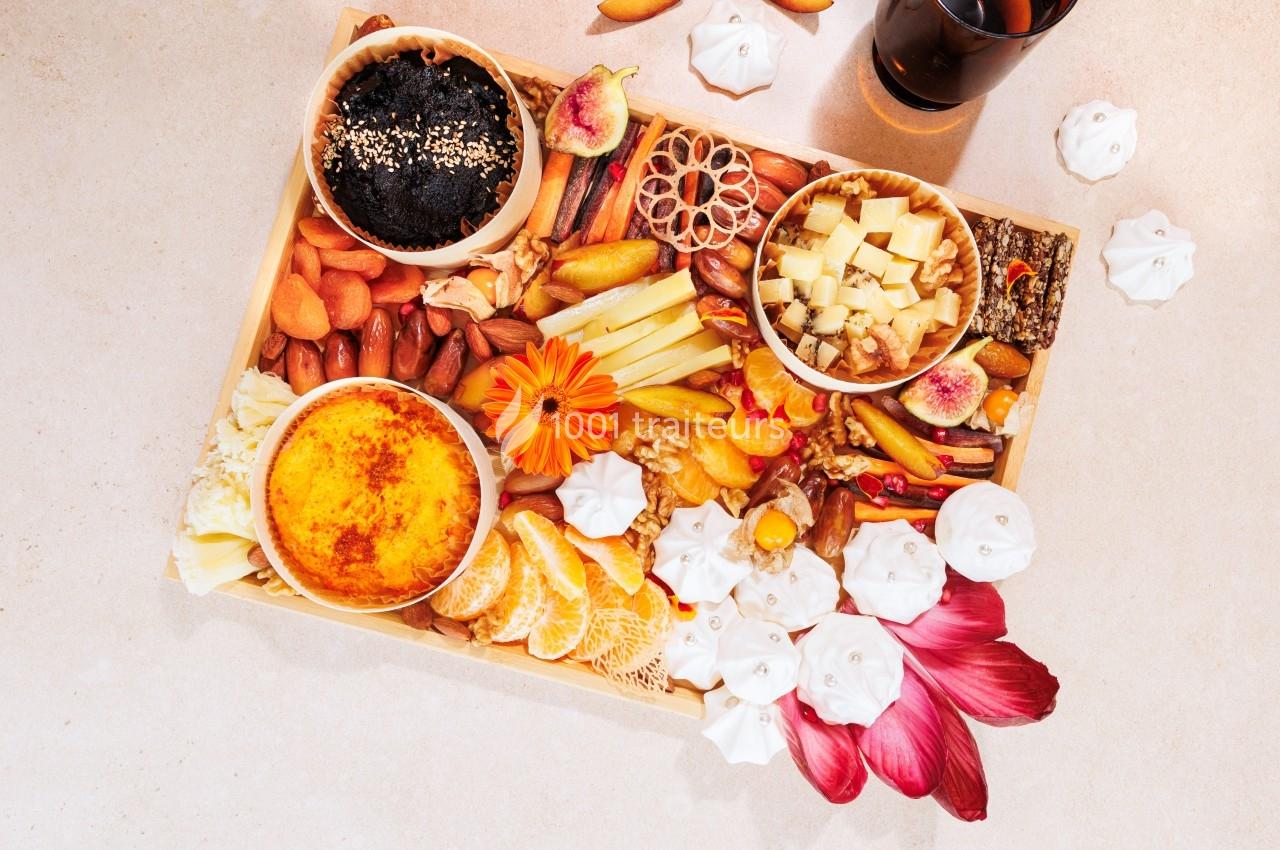 Plateau varié avec fruits, fromages, noix, charcuteries, crème brûlée et décorations florales sur une table claire.