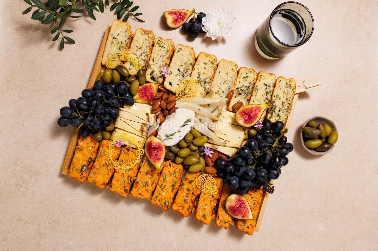 Plateau garni de fromages, fruits frais, olives, noix et tranches de pain, disposé sur une table beige.