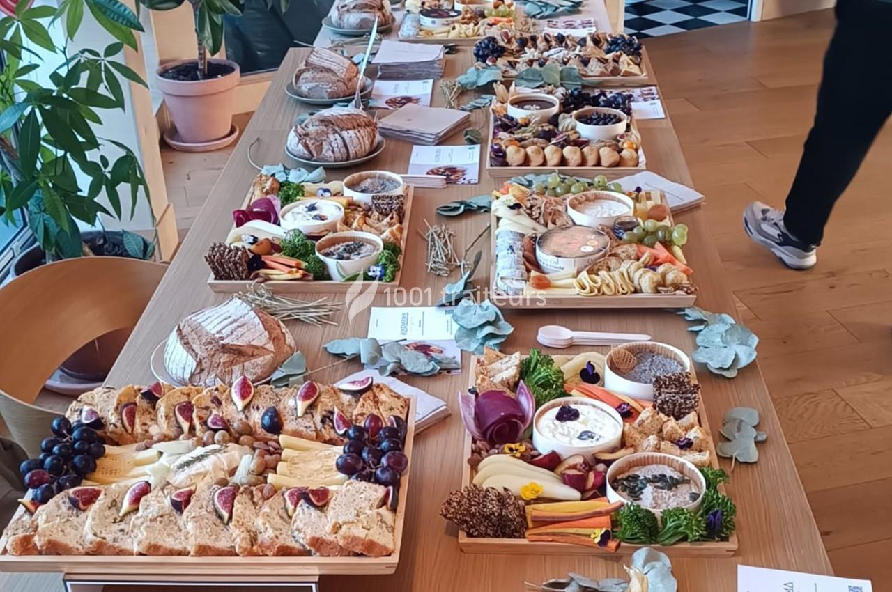 Table garnie de plateaux de charcuterie, fromages, fruits et pains, disposés pour un buffet dans un espace lumineux.