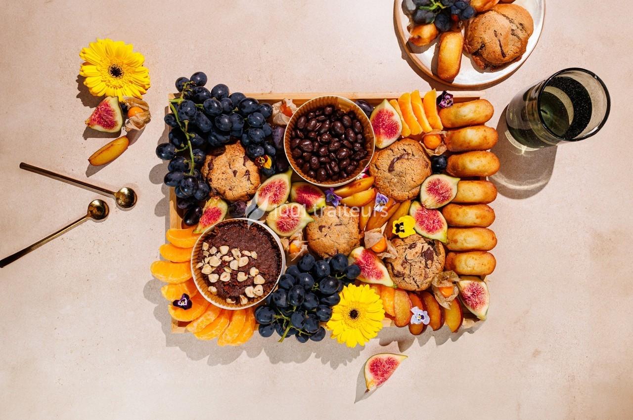 Plateau garni de fruits frais, muffins, biscuits, chocolats et fleurs décoratives sur une table beige.