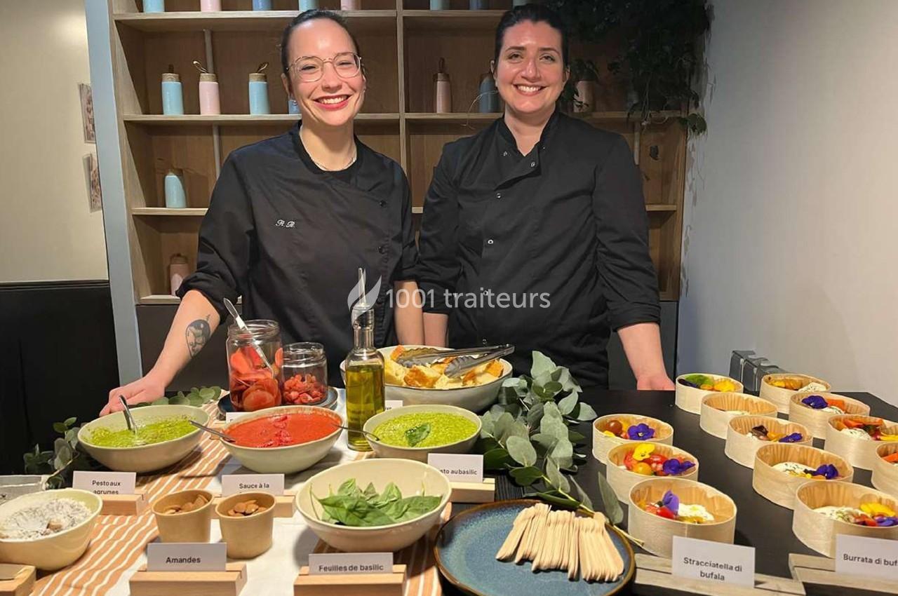 Deux cheffes souriantes derrière une table garnie de plats colorés, sauces, légumes et garnitures variées.