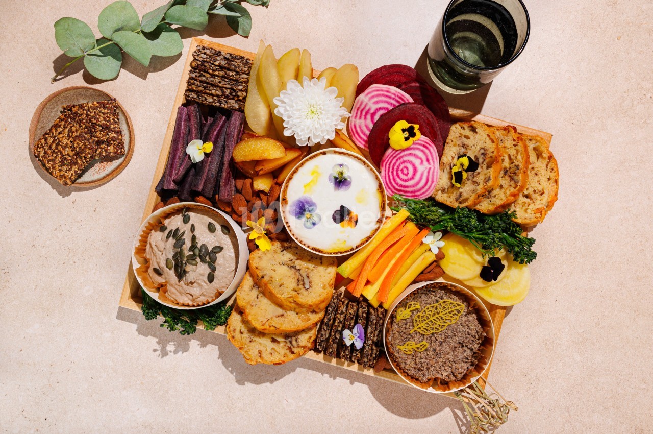 Plateau de dégustation avec légumes, tartinades, crackers, fruits et fleurs comestibles, disposé sur une table beige.