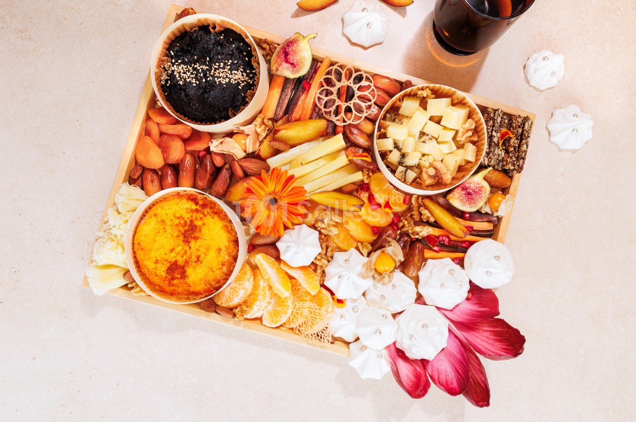 Plateau varié avec fruits, fromages, noix, charcuteries, crème brûlée et décorations florales sur une table claire.