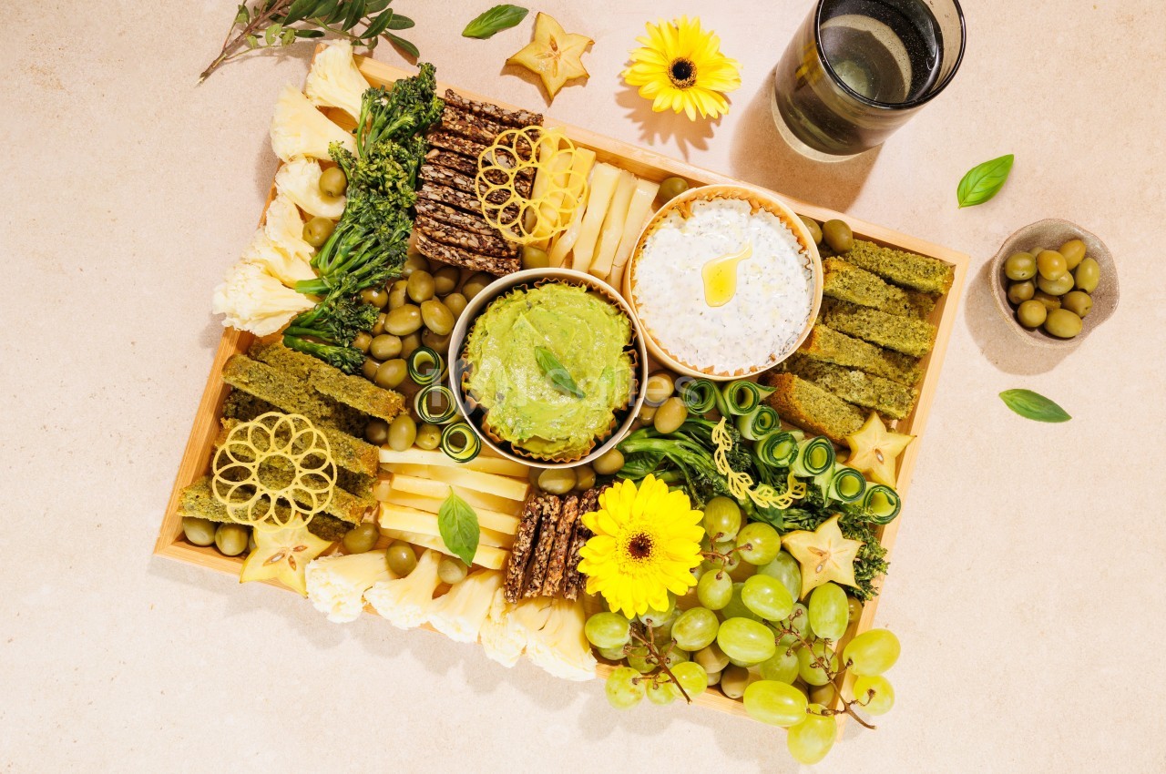 Plateau de dégustation avec fruits, légumes, fromages, crackers et sauces, décoré de fleurs jaunes.